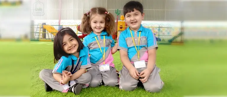 Three young children in school uniform sitting together on the grass beside a kindergarten play area